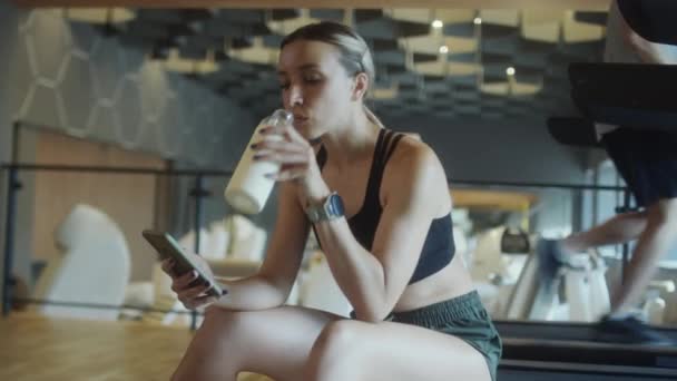 スポーツウェアを着た女性がジムに座り、ボトルから水を飲み、休憩中に携帯電話を使用して背景にトレッドミルでジョギングをしています - 映像、動画