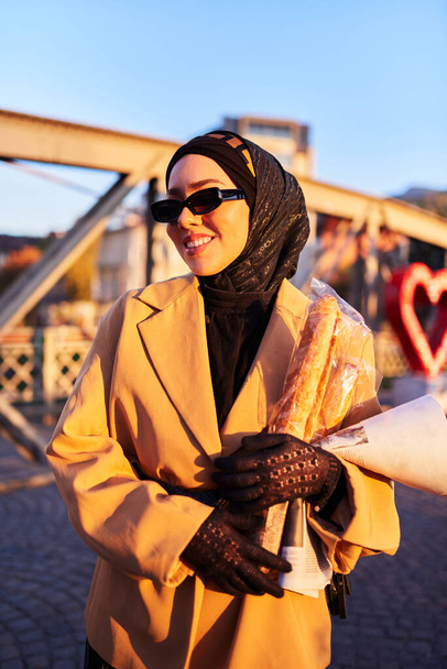 Женщина в хиджабе в стильных солнцезащитных очках и элегантном французском наряде, прогуливаясь по городу на закате солнца, неся букет, хлеб и газету, излучая чувство культурного шарма и спокойствия.  - Фото, изображение