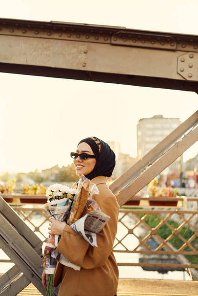 Жінка з хіджабу в стильних сонцезахисних окулярах і елегантний французький одяг, прогулюючись по місту під час заходу сонця, несучи букет, хліб і газету, випромінюючи почуття культурної чарівності і спокою..  - Фото, зображення