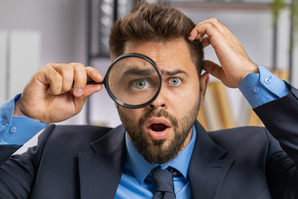 Investigator researcher scientist business man working at office holding magnifying glass near face looking into camera with big zoomed funny eye, searching, analysing. Professional freelancer guy - Photo, Image