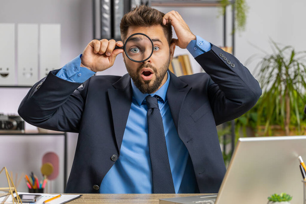 Investigator researcher scientist businessman working at home office holding magnifying glass near face looking into camera with big zoomed funny eye, searching, analysing. Professional freelancer man - Foto, afbeelding