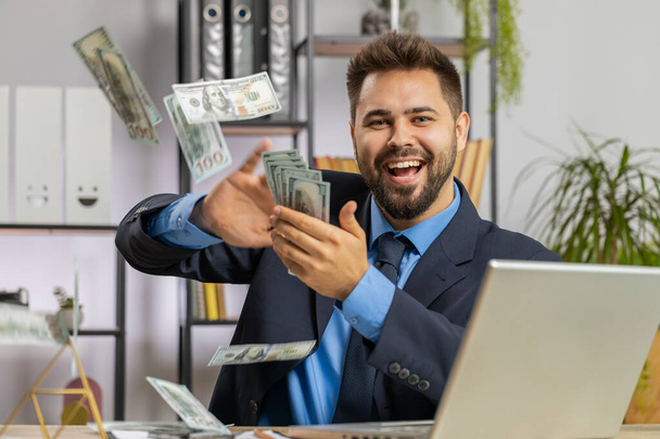 Cheerful rich Caucasian businessman boss working on laptop and wasting throwing money to camera more tips earnings big profit success win lottery shopping. Happy freelancer man guy at home office desk - Foto, Imagem