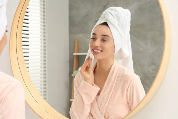 Beautiful woman in terry towel removing makeup with cotton pad near mirror indoors - 写真・画像