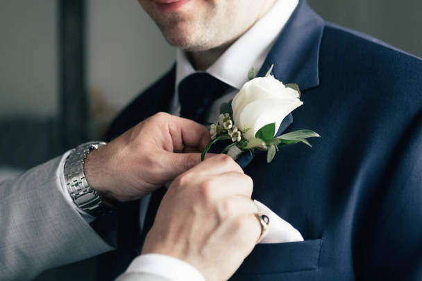 Damadın gül yaka çiçeğini takmasına yardım eden bir arkadaşın yakın çekimi. Damat, düğün töreni için hazırlanıyor ve otel odasında giyiniyor. Damadın arkadaşına yardım et. Gül yaka çiçeği aksesuarı - Fotoğraf, Görsel