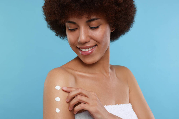 Beautiful young woman applying body cream onto arm on light blue background - Фото, изображение