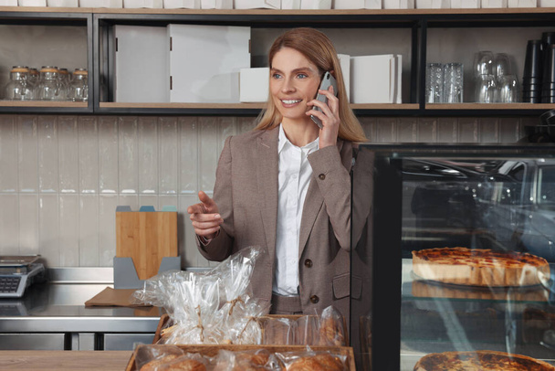 Happy business owner talking on phone at cashier desk in bakery shop - Фото, изображение