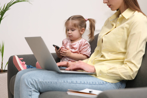 Mother working remotely on laptop while her daughter playing with smartphone at home, closeup - 写真・画像