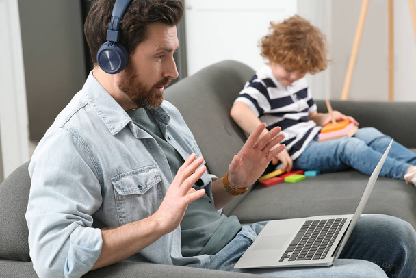 Father working remotely on laptop while his son playing with toys at home. Man having video chat with colleagues - 写真・画像