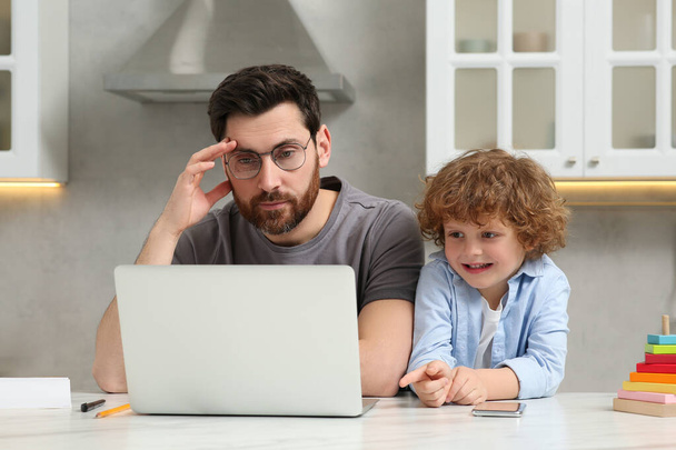 Little boy bothering father while he working remotely at home. Man with laptop and his child at desk in kitchen - 写真・画像