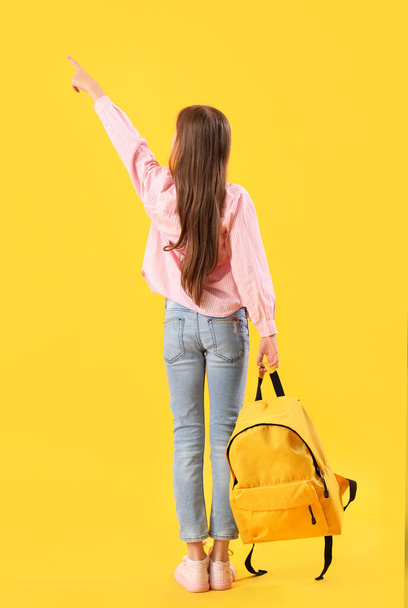 Niña con mochila apuntando a algo sobre fondo amarillo, vista trasera - Foto, Imagen