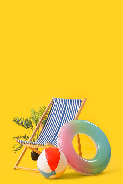 Deck chair with beach ball, inflatable ring and palm tree on yellow background - Fotó, kép