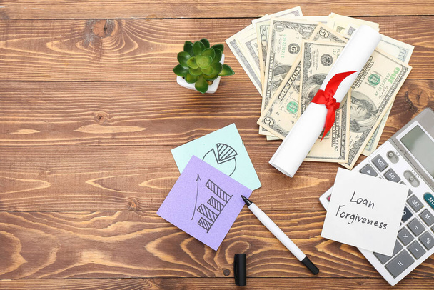 Diploma, dollar banknotes, calculator and sticky note with text LOAN FORGIVENESS on wooden background - 写真・画像