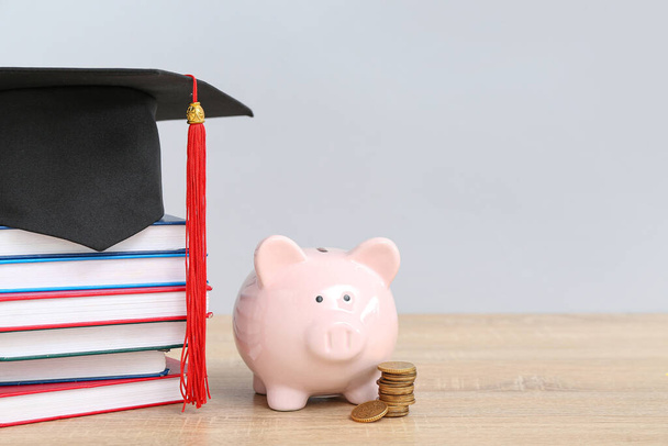 Свинка банка, стопка монет, книги и выпускной шляпы на деревянном столе на сером фоне. Концепция студенческого кредита - Фото, изображение