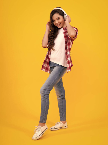 Teenager in modern wireless headphones enjoying song music in audio app, listening to her favorite music on yellow background. Happy teenager girl casual wear. Urban kids style, stylish hipster teen - 写真・画像