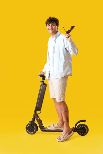 Joven con gafas de sol y scooter eléctrico sobre fondo amarillo - Foto, Imagen
