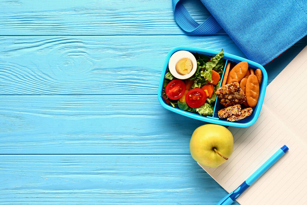 Rugzak, briefpapier en lunchbox met lekker eten op blauwe houten ondergrond - Foto, afbeelding