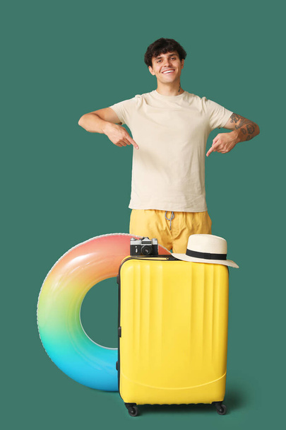Junger Mann zeigt auf Koffer mit Kamera und Hut auf grünem Hintergrund - Foto, Bild