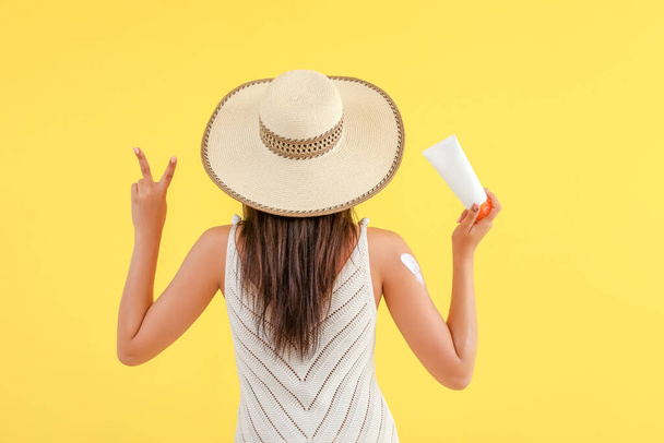 Beautiful woman with sunscreen cream showing victory gesture on yellow background, back view - Photo, Image
