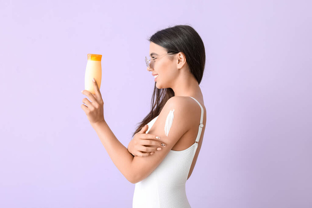 Mooie jonge vrouw met zonnebrandcrème op lila achtergrond - Foto, afbeelding