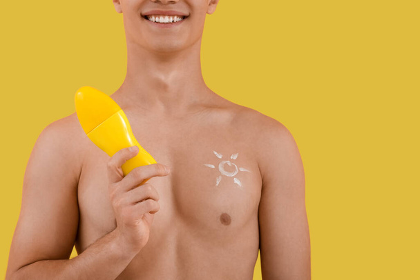 Happy young man with bottle of sunscreen cream on yellow background, closeup - Photo, Image
