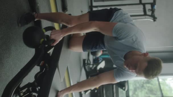 Tiro vertical de hombre musculoso en ropa deportiva haciendo Kettlebell ejercicio de fila de un solo brazo en el banco mientras entrena en el gimnasio - Imágenes, Vídeo