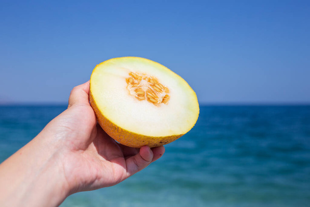 hand holding a piece of ripe watermelon on a background of a blue sea and a white sand beach, close - up, copy space, top - Foto, Imagen