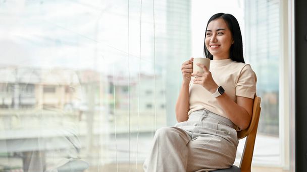 Μια όμορφη και ευτυχισμένη Ασιάτισσα με καθημερινά ρούχα που απολαμβάνει τον πρωινό της καφέ σε μια καρέκλα δίπλα στο παράθυρο.. - Φωτογραφία, εικόνα