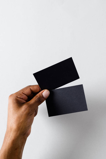 Vertical image of hand of biracial man with black business cards with copy space on white background. Business, business card, stationery and writing space concept. - Zdjęcie, obraz