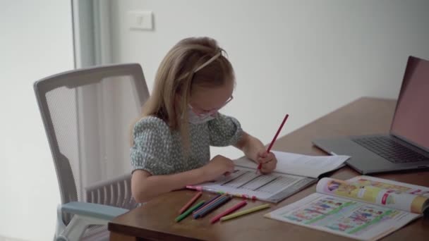 Pieni fiksu söpö tyttö alakouluikäisissä laseissa tekee englannin läksyjä suojaavassa naamiossa. Käsite lasten peruskoulutuksesta. Lapsi katsoo tunteja kotona. Pandemia - Materiaali, video