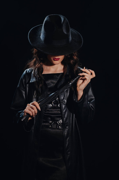 Sexy female dominant mistress with a whip for BDSM sex with submission and domination in a leather raincoat and a hat in the noir style - Фото, изображение