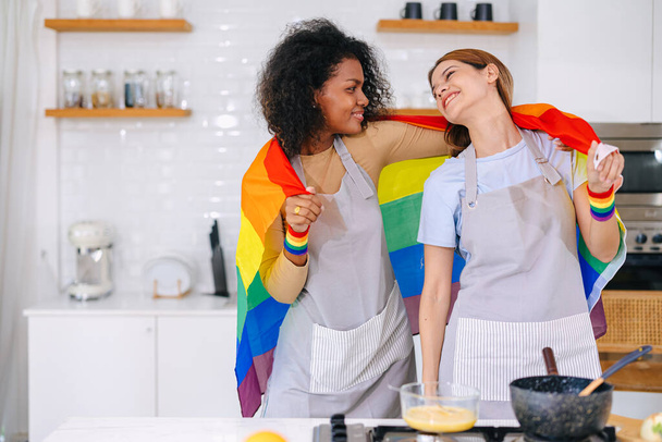 ΛΟΑΤ ζευγάρι γυναίκες ευτυχείς γιορτάζουν μήνα υπερηφάνειας μαζί με τη σημαία ουράνιο τόξο στην κουζίνα μαγείρεμα στο σπίτι - Φωτογραφία, εικόνα