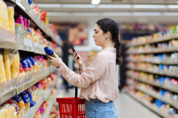Side view of caucasian woman scan qr code of product using smartphone. Shelves with food in background. Concept of shopping and consumerism. - 写真・画像