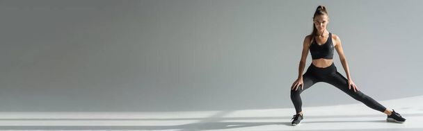 брюнетка спортсменка в спортивном бюстгальтере и леггинсах, смотрящая в камеру, упражняясь на сером фоне  - Фото, изображение