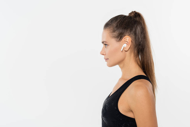 Boční pohled sportovkyně v černém sportovním oblečení pomocí bezdrátového sluchátka izolované na bílém - Fotografie, Obrázek