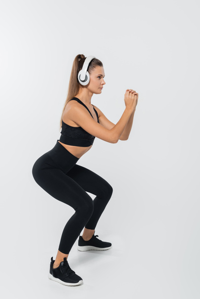sportvrouw in hoofdtelefoon en sportkleding hurken op witte achtergrond, wellness-concept - Foto, afbeelding