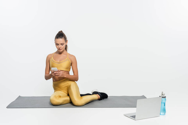 mulher usando smartphone e sentado o tapete de fitness perto de laptop em fundo branco, atividade física - Foto, Imagem