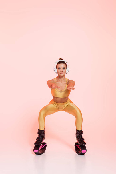 resistencia y equilibrio, mujer en zapatos de salto de kangoo y auriculares inalámbricos haciendo sentadillas en rosa - Foto, imagen