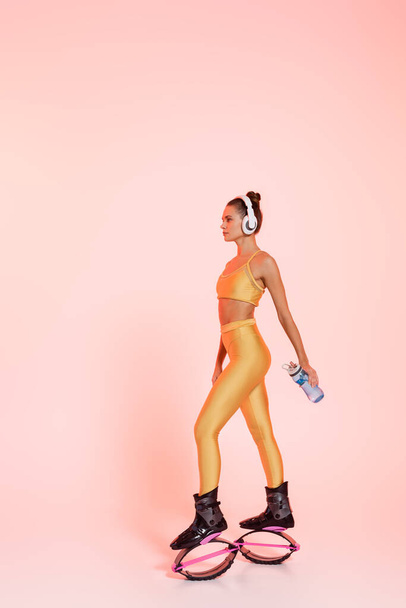 woman in kangoo jumping shoes and wireless headphones holding sports bottle with water on pink  - Photo, Image
