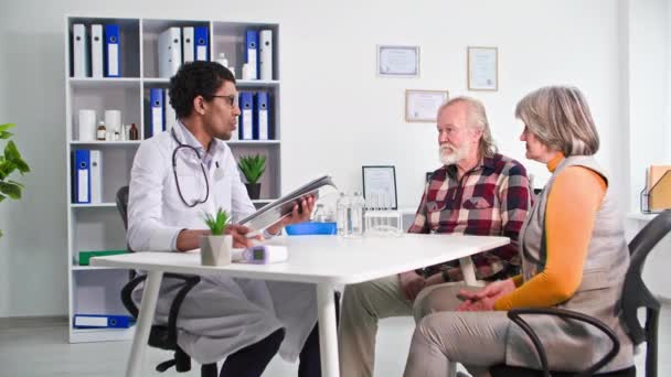 medical consultation, black doctor examines an elderly couple in hospital office - Materiał filmowy, wideo