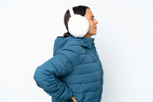 Молодая испаноязычная женщина в зимних наушниках, изолированных на белом фоне, страдает от боли в спине за то, что сделала усилие - Фото, изображение