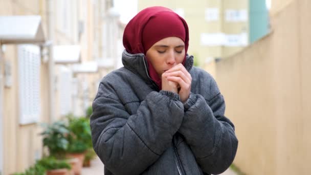 Мусульманська молода жінка в хіджабі і пальто розігріває руки з диханням, кашляє і дме їй ніс в тканині на вулиці. - Кадри, відео