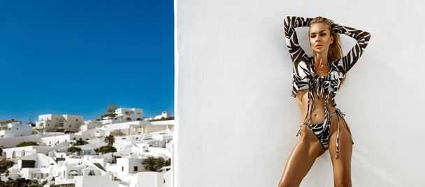Elegante modelo rubia sexy en bikini está posando sensualmente en un resort de lujo con vista a la isla de Santorini. - Foto, Imagen