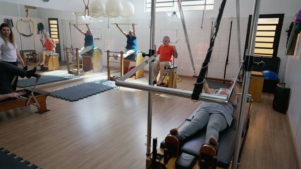 Pilates Stüdyosunda egzersiz yapan bir grup yaşlı insan. Yaşlılık egzersizleri. Yaşlılara vücut geliştirme ve esnetme eğitimi veren kadın antrenör - Fotoğraf, Görsel