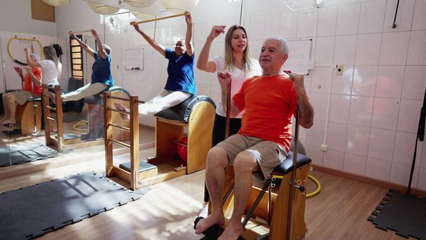 Nauczyciel pilatesu trenuje seniora z maszyną. Grupowa sesja fizjoterapeutyczna, rutynowy trening starości - Zdjęcie, obraz