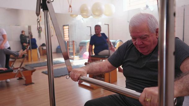 Starsza osoba ćwicząca w Pilates Studio Group Session. Jeden starszy mężczyzna rasy kaukaskiej dbający o ciało za pomocą maszyny - Zdjęcie, obraz