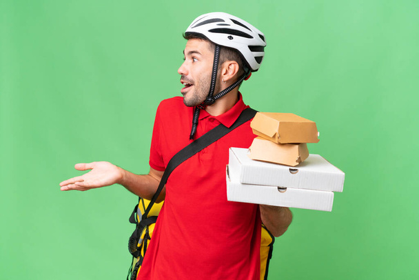 Young handsome caucasian man with thermal backpack and holding takeaway food over isolated background with surprise expression while looking side - Photo, Image