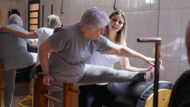 女性ピラティスコーチは理学療法のクラスで体を伸ばすためにシニア女性を導く。古い時代のワークアウト｜ルーチンの健康と健康ライフスタイル - 写真・画像