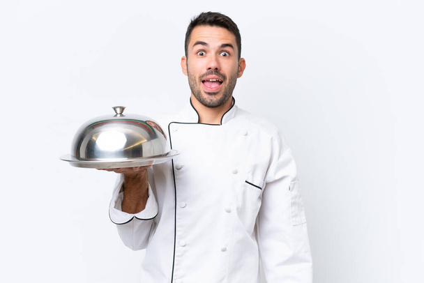 Joven chef con bandeja aislada sobre fondo blanco con expresión facial sorpresa - Foto, imagen