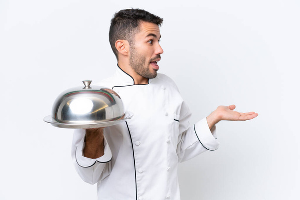 Молодий шеф-кухар з підносом ізольований на білому тлі з несподіваним виразом під час огляду сторони - Фото, зображення