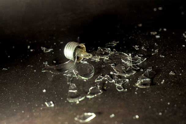 A broken light bulb, filament dangling and glass shattered, with jagged shards laid around what remains of the bulb - Foto, Bild